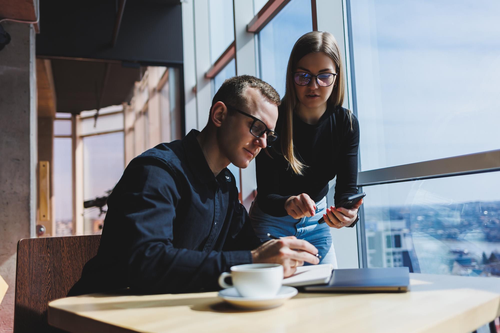 Two colleagues at a café table look over a laptop and a phone.