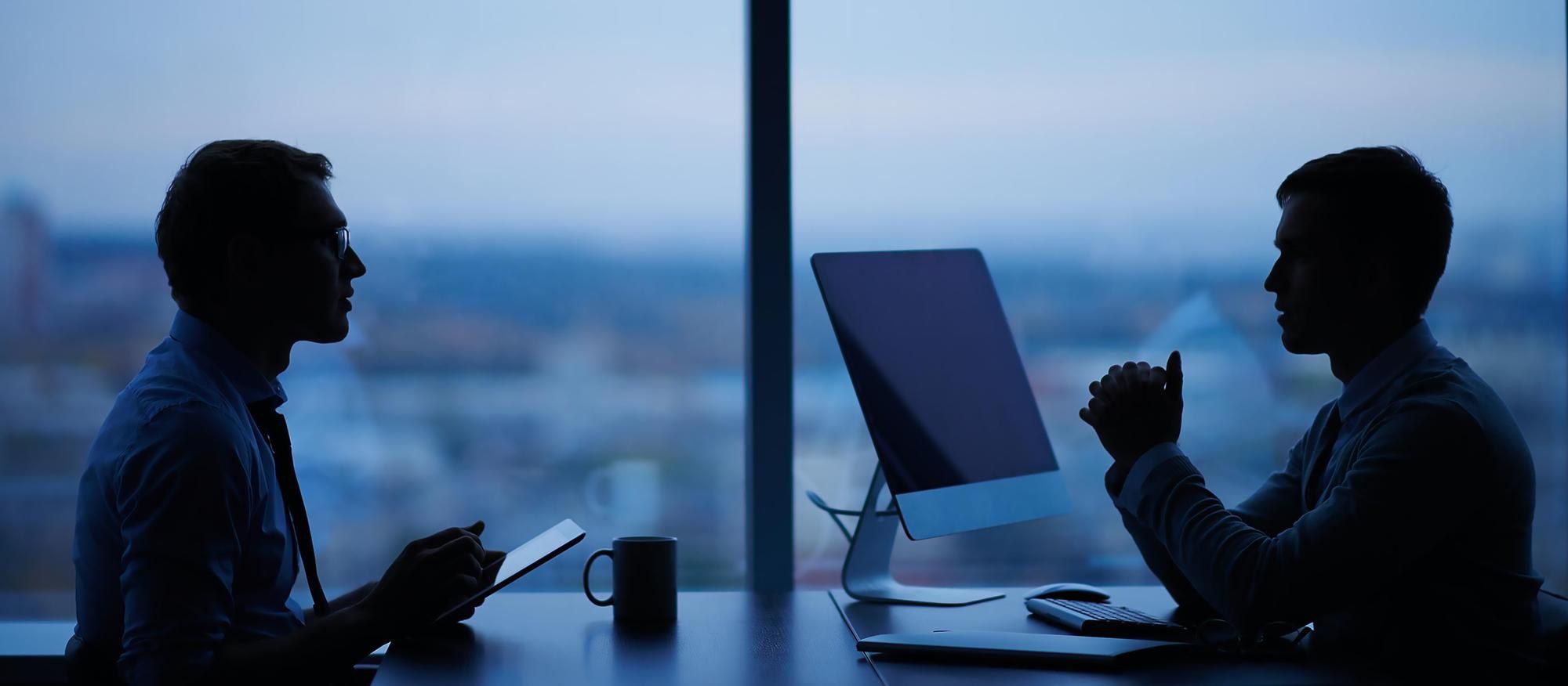 Two colleagues work late at a desk by a window overlooking the city.